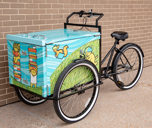 The book bike looks like a tricycle with the double wheels in the front, flanking a blue and green painted box with characters holding books and wearing helmets. The book bike looks like a tricycle with the double wheels in the front, flanking a blue and green painted box with characters holding books and wearing helmets.