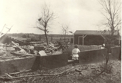 black and white photograph of a destroyed and collapsed home. A child looks at the destruction.