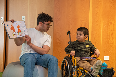 A man reading a book that is in English and Spanish. He is smiling at a young boy in a wheelchair.