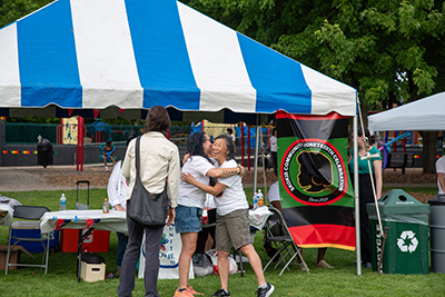 Two people are embracing outside of an outdoor event tent. There is a flag of a Black person's silhouette in the colors black and red hanging from the tent