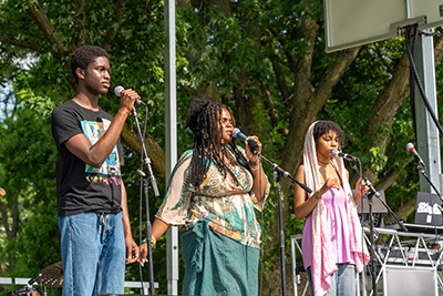 Three people standing on an outdoor stage and singing into microphones