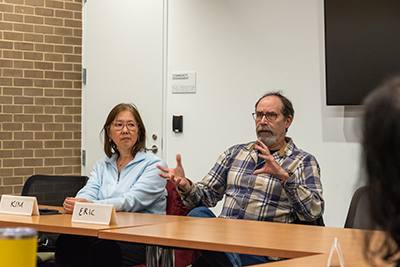 Two people sitting at a table next to each other. One person is gesturing and speaking" title="Two people sitting at a table next to each other. One person is gesturing and speaking