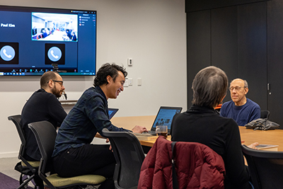 Four people sitting across from each other at a table. One person has a laptop and there are zoom participants on a screen on the wall " title="four people sitting across from each other at a table. One person has a laptop and there are zoom participants on a screen on the wall