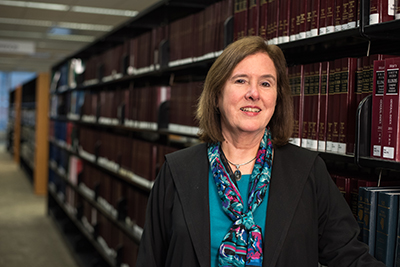A smiling woman in a teal shirt and teal/purple scarf standing in front of a bookcase in the library