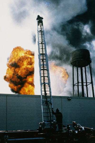 Firefighter on top of a tall ladder above a large fire. Water tower in the background.