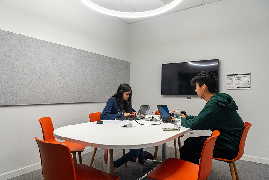Two teens, one girl and one boy, sitting opposite the other in a study room with open laptops on the table