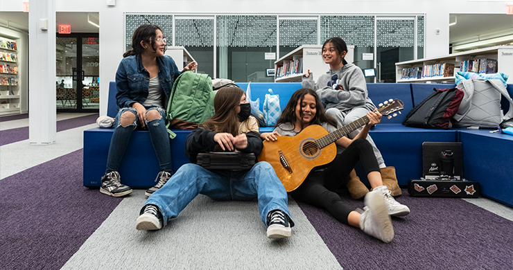 Group of four teen girls sitting in the library. Two are mid-conversation while one holds a guitar