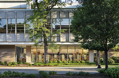 Multi story building with glass windows across the front on each level. Three trees in front of the building and lots of greenery.