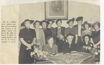 Old black and white newspaper clipping of women in fancy clothes and hats standing behind three other women sitting at a table.
