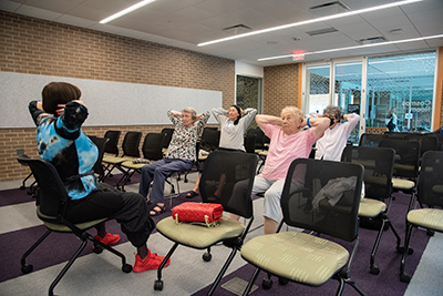 Rows of chairs lined up with four ladies sitting and doing exercising looking at the lady in the front leading the exercises.