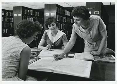 Black and white photo of three women at a table in the library looking at large books