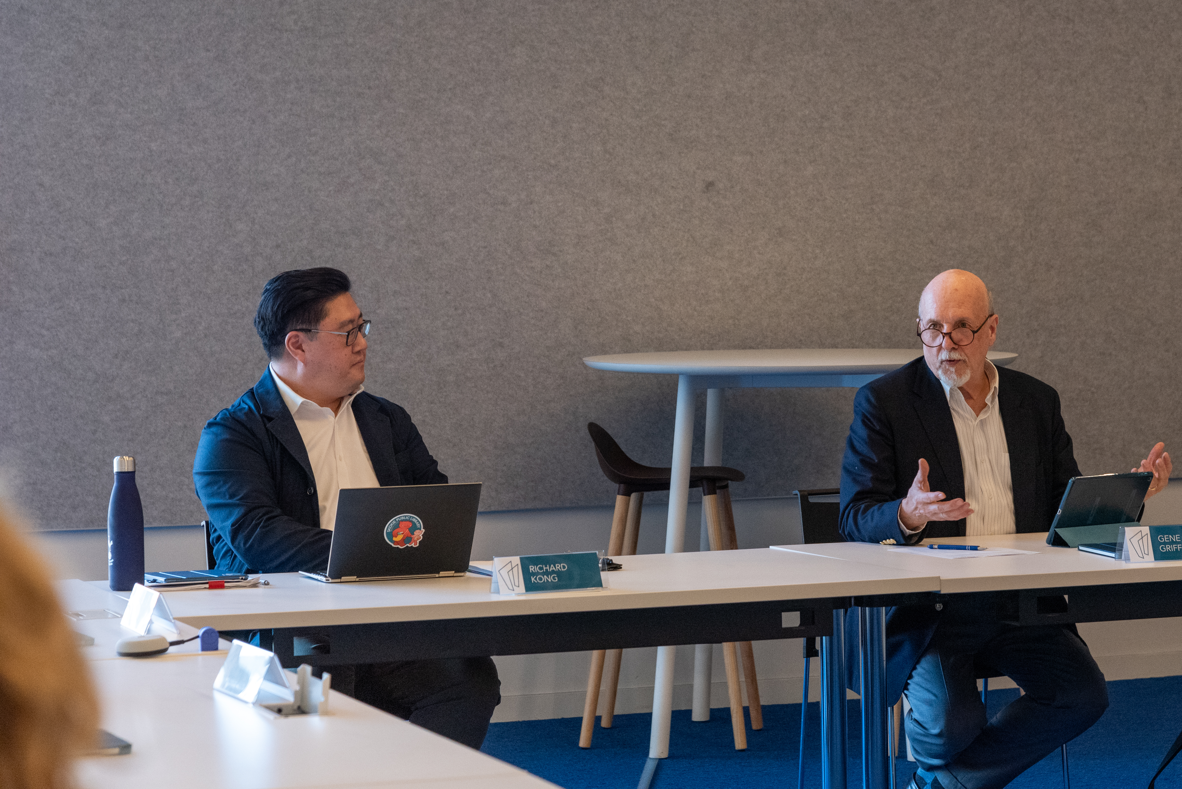 Richard Kong and Gene Griffin sitting around a table. Gene is gesturing and speaking.