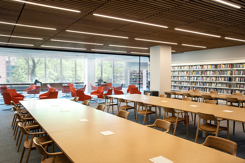 The current Reading Room in the Skokie Public Library, showing long wooden tables with wooden chairs, bookshelf-lined walls, and low orange chairs along a floor to ceiling wall of glass that looks out on the second floor of the library." title="The current Reading Room in the Skokie Public Library, showing long wooden tables with wooden chairs, bookshelf-lined walls, and low orange chairs along a floor to ceiling wall of glass that looks out on the second floor of the library.