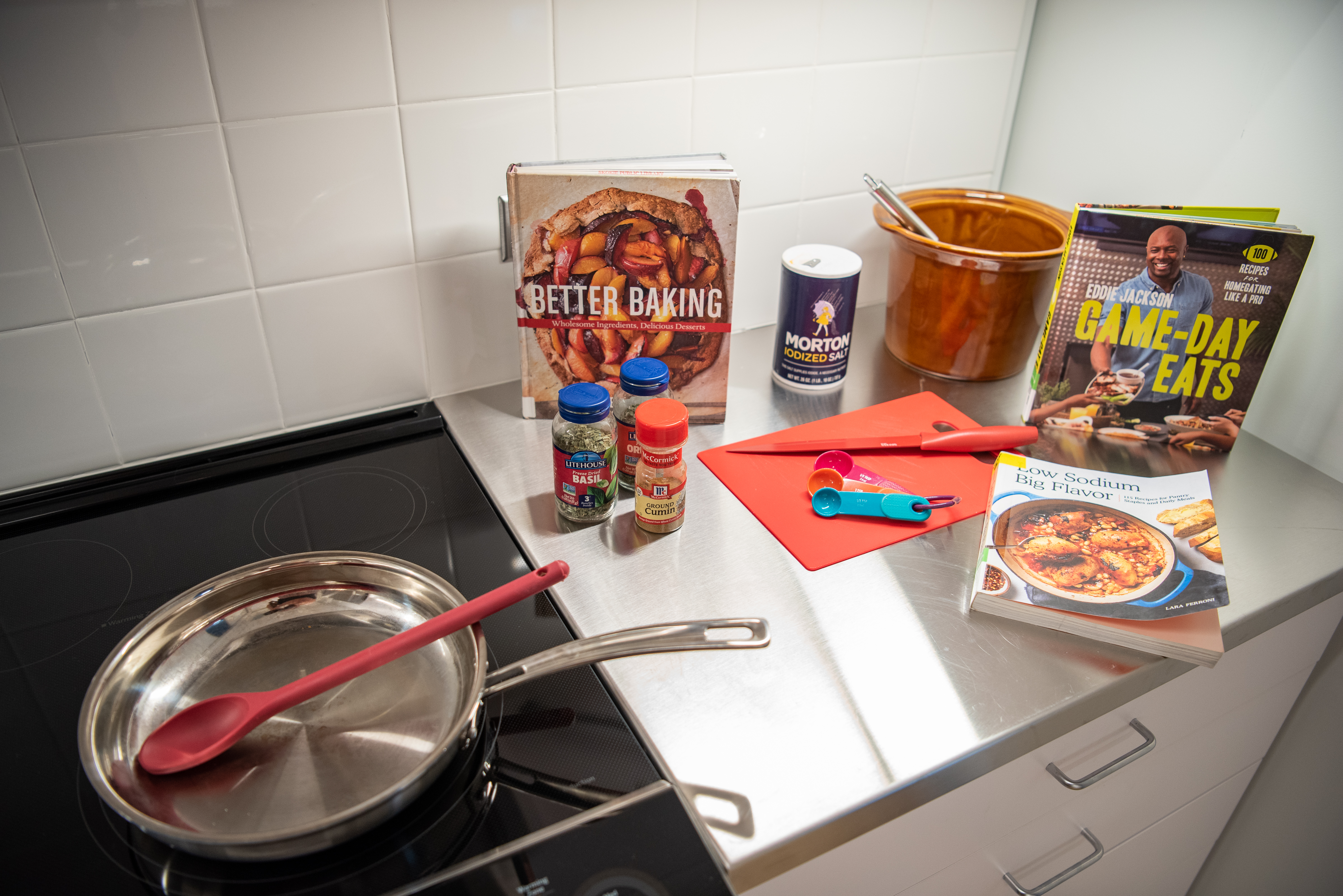 three cookbooks on a counter next to a cutting board, knife, spices, and pan