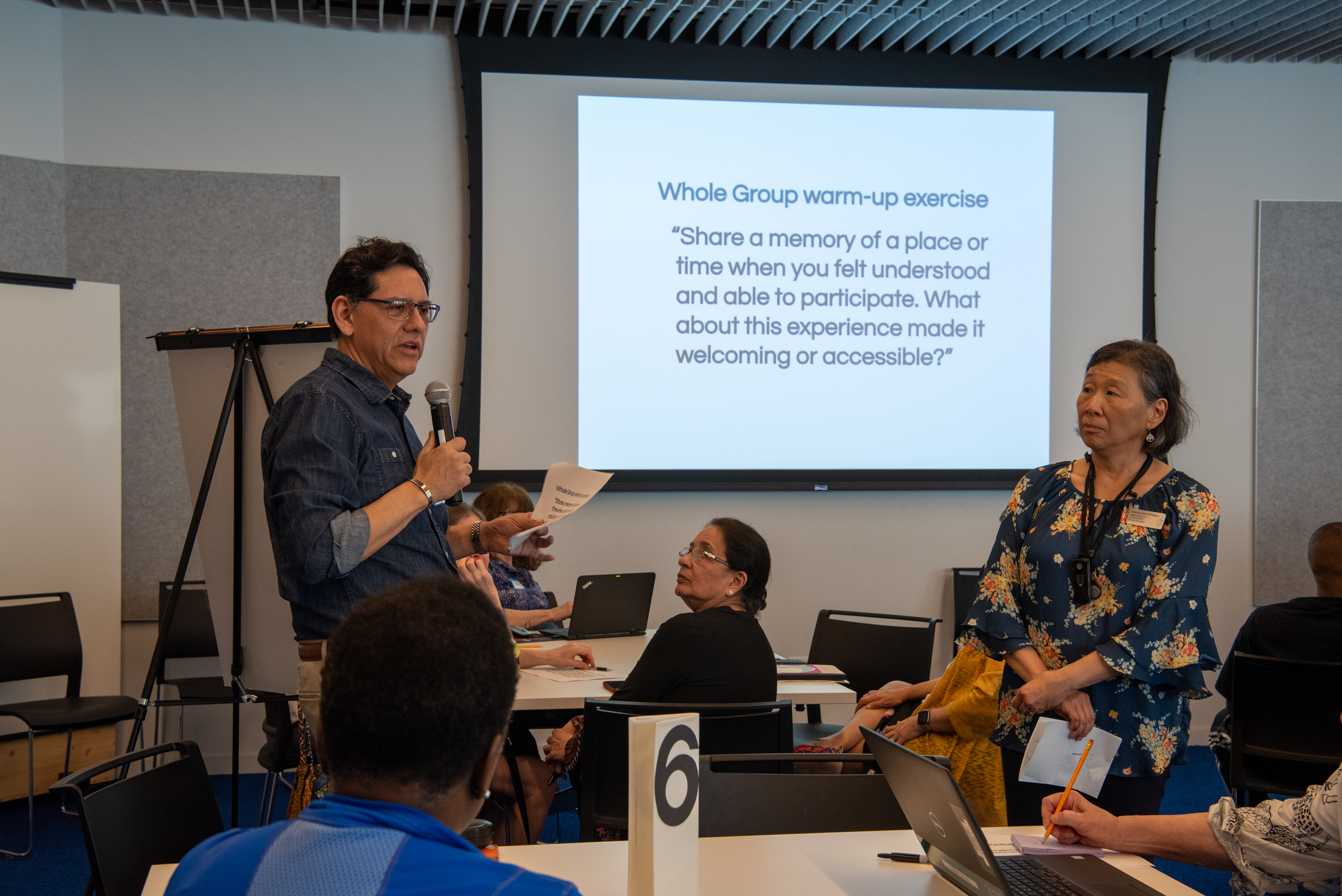 person holding a mic and talking to an audience. Projection on the wall reads "share a memory of aplace or time when you felt understood and able to participate. What about his experience made it welcoming or accessible?"