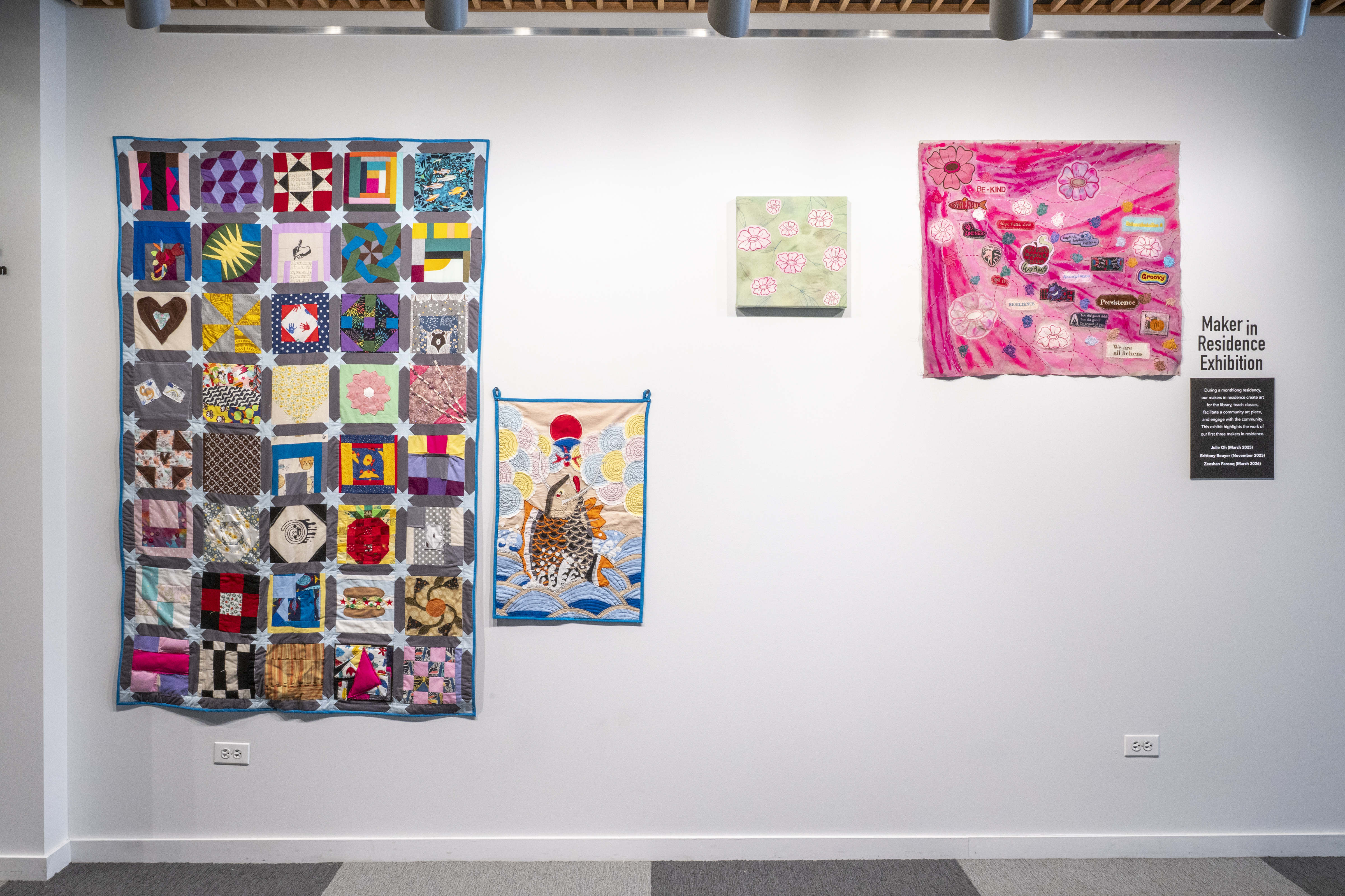 a wall with vinyl text that reads "Maker in Residence Exhibition" There are four fabric art pieces hanging on the wall. 