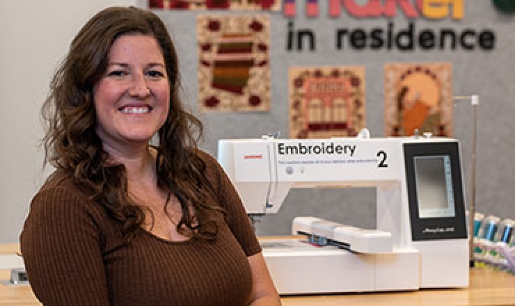 a woman smiling in front of a work table with an embroidery machine on it. 