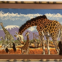 Needlepoint tapestry depicting a herd of giraffes with mountains and sky in the background.