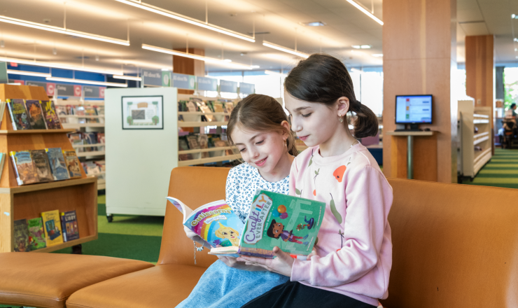 Two young sisters sitting side-by-side in the library holding open books