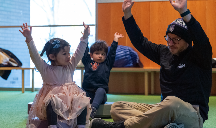 Man sitting with two children, all three raising their hands during a library storytime event